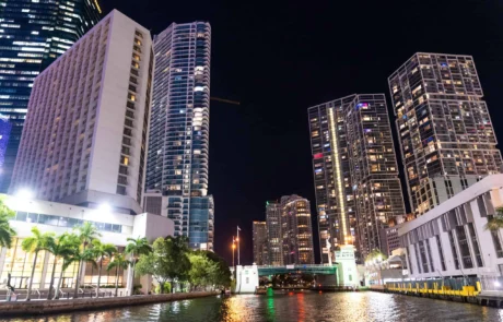 Nighttime view of a Miami waterfront with illuminated skyscrapers and modern buildings, reflecting on the water, alongside a raised drawbridge. Nighttime view of a Miami waterfront with illuminated skyscrapers and modern buildings, reflecting on the water, alongside a raised drawbridge.