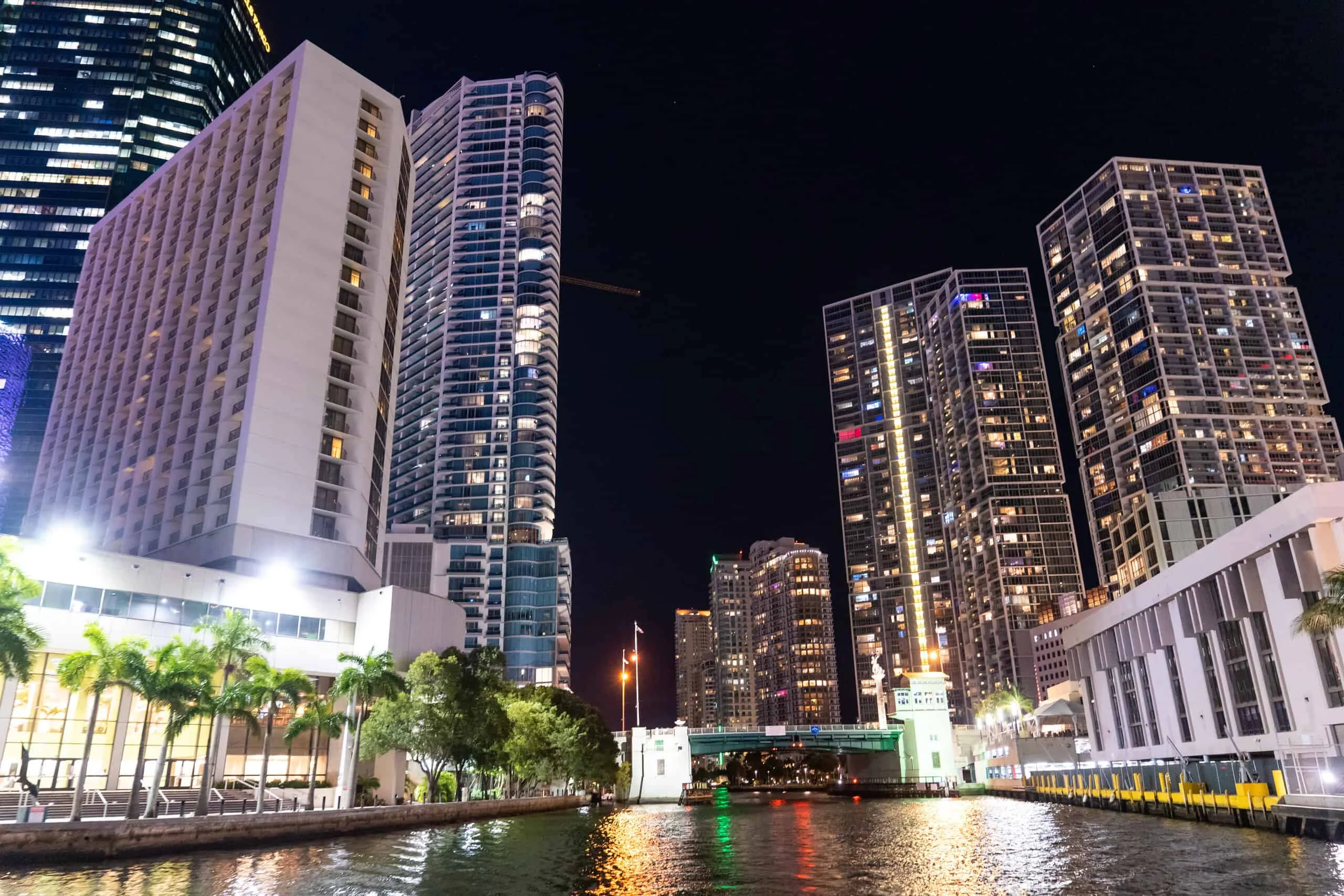 Nighttime view of a Miami waterfront with illuminated skyscrapers and modern buildings, reflecting on the water, alongside a raised drawbridge. Nighttime view of a Miami waterfront with illuminated skyscrapers and modern buildings, reflecting on the water, alongside a raised drawbridge.