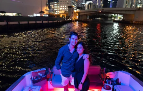 A couple smiling and standing together on a boat in Miami with illuminated city buildings in the background at night.