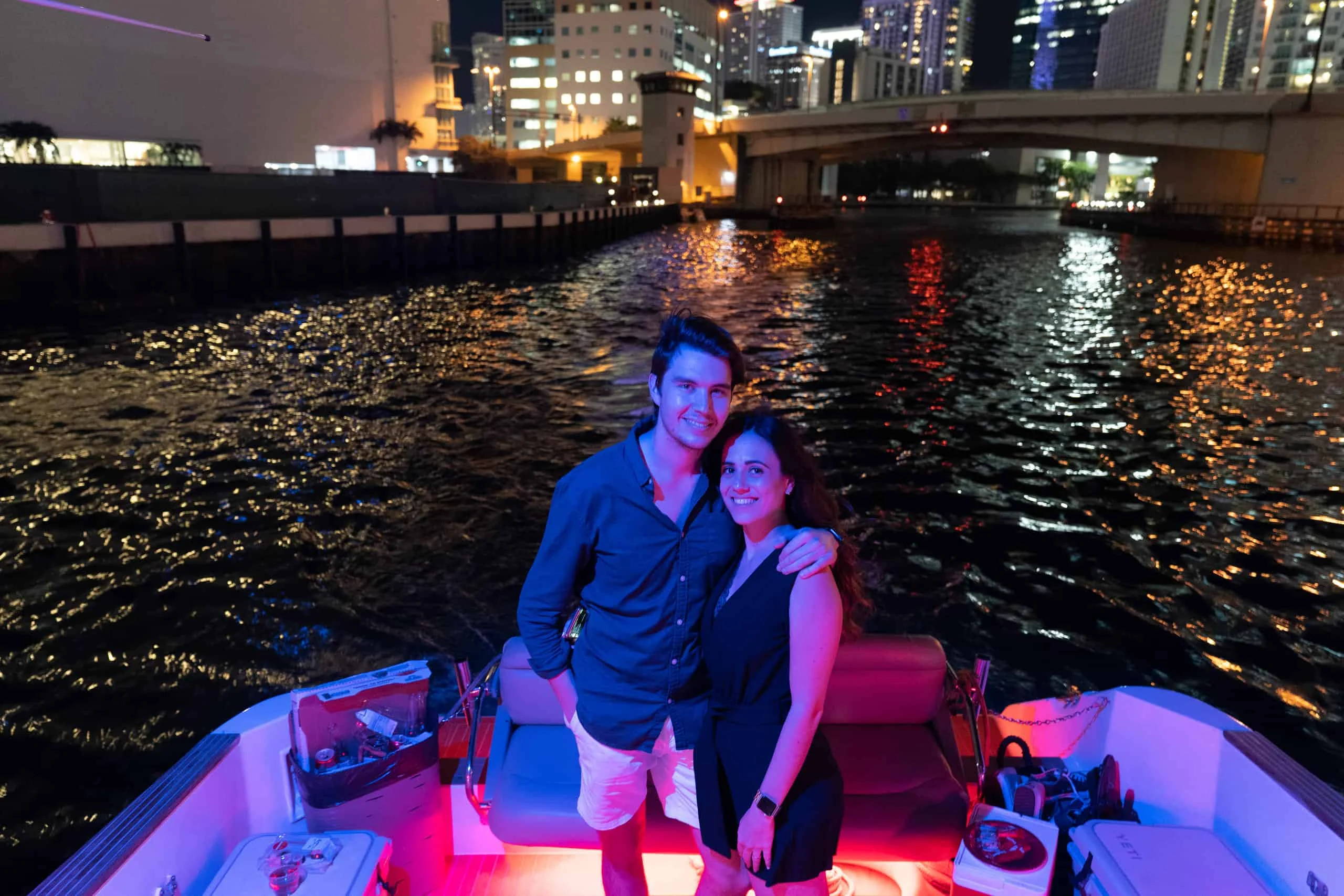 A couple smiling and standing together on a boat in Miami with illuminated city buildings in the background at night. A couple smiling and standing together on a boat in Miami with illuminated city buildings in the background at night.