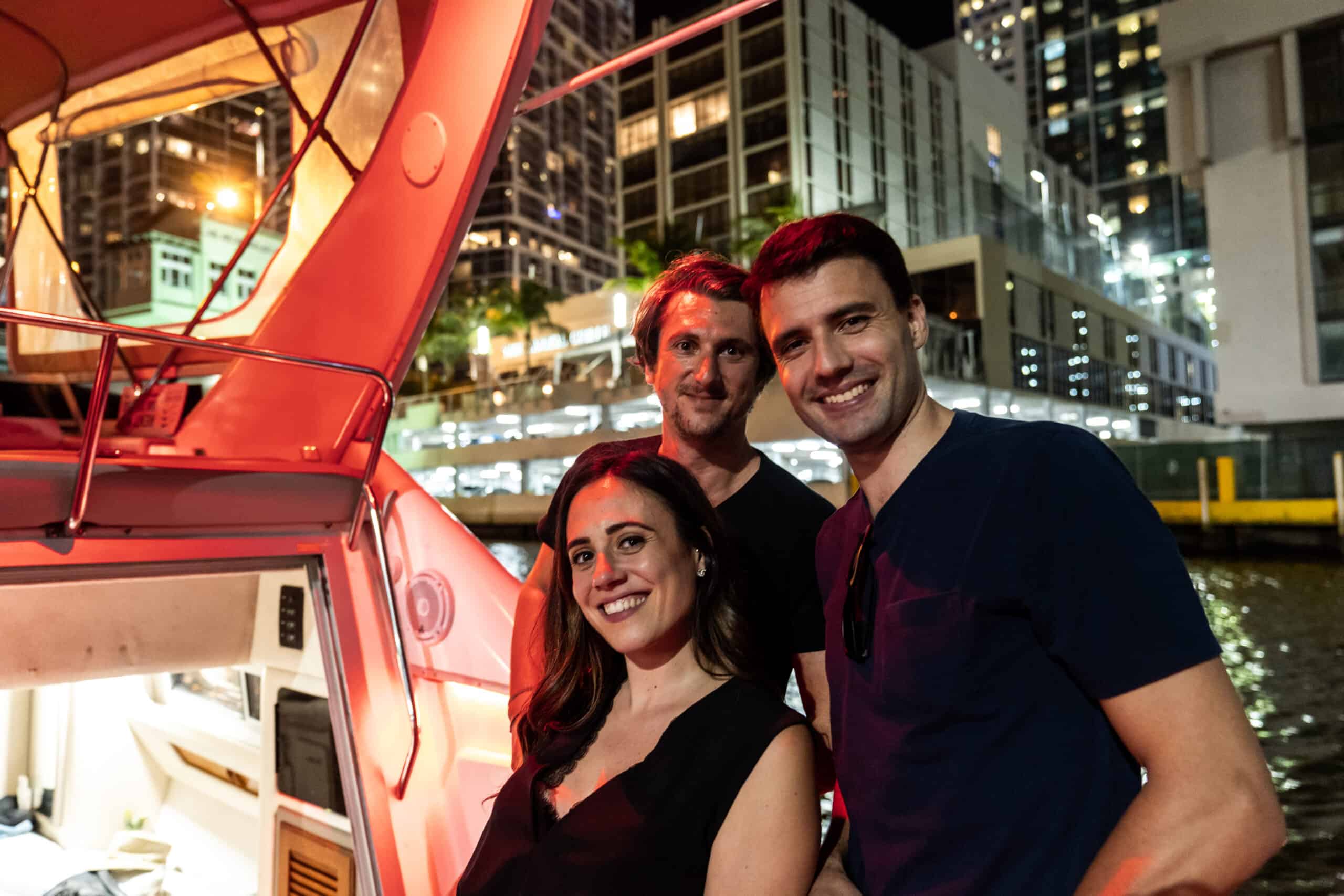 Three friends smiling on a boat in Miami at night with illuminated city buildings in the background.