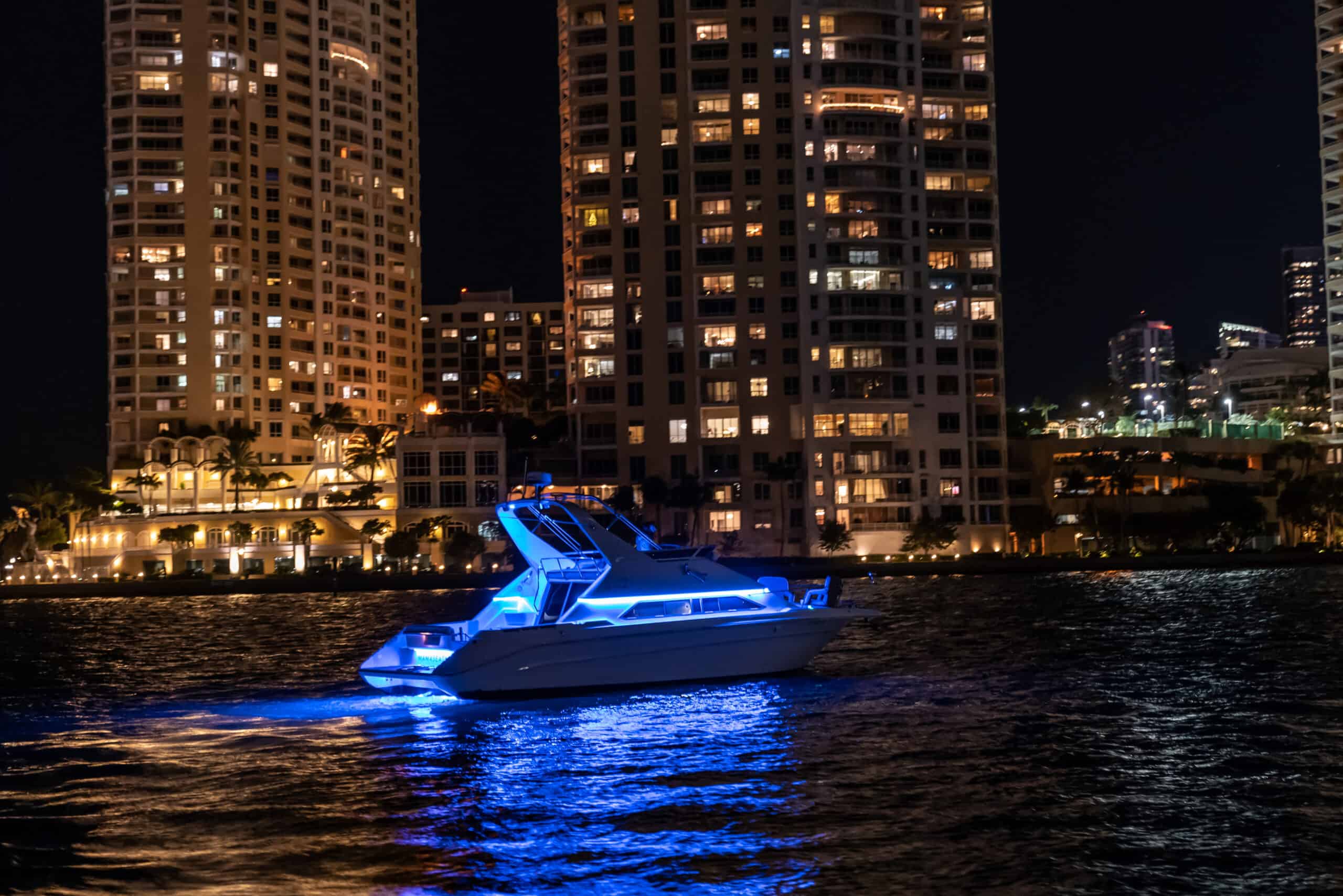 A yacht illuminated with blue lights floating on water at night, with brightly lit high-rise buildings in Miami in the background.