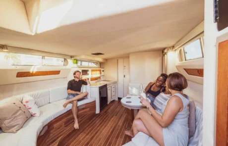 Three people enjoying conversation in a cozy, well-lit yacht cabin with wooden floors and white furniture, sailing off the coast of Miami.