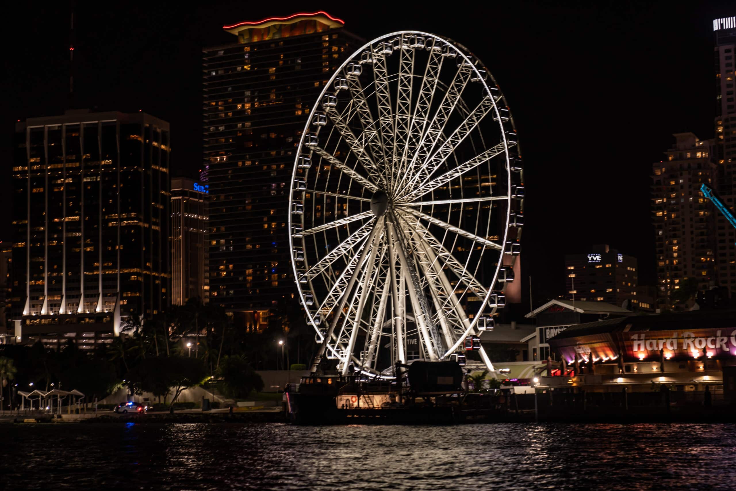 Nighttime cityscape with a lit ferris wheel near waterfront buildings, including a hard rock cafe, against a backdrop of illuminated high-rises in Miami.