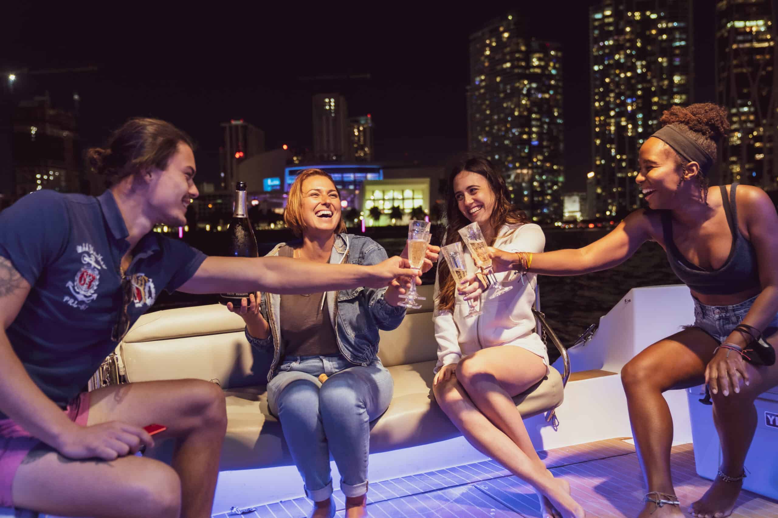 Four friends, holding drinks, joyfully toast on a boat at night with Miami city lights in the background.