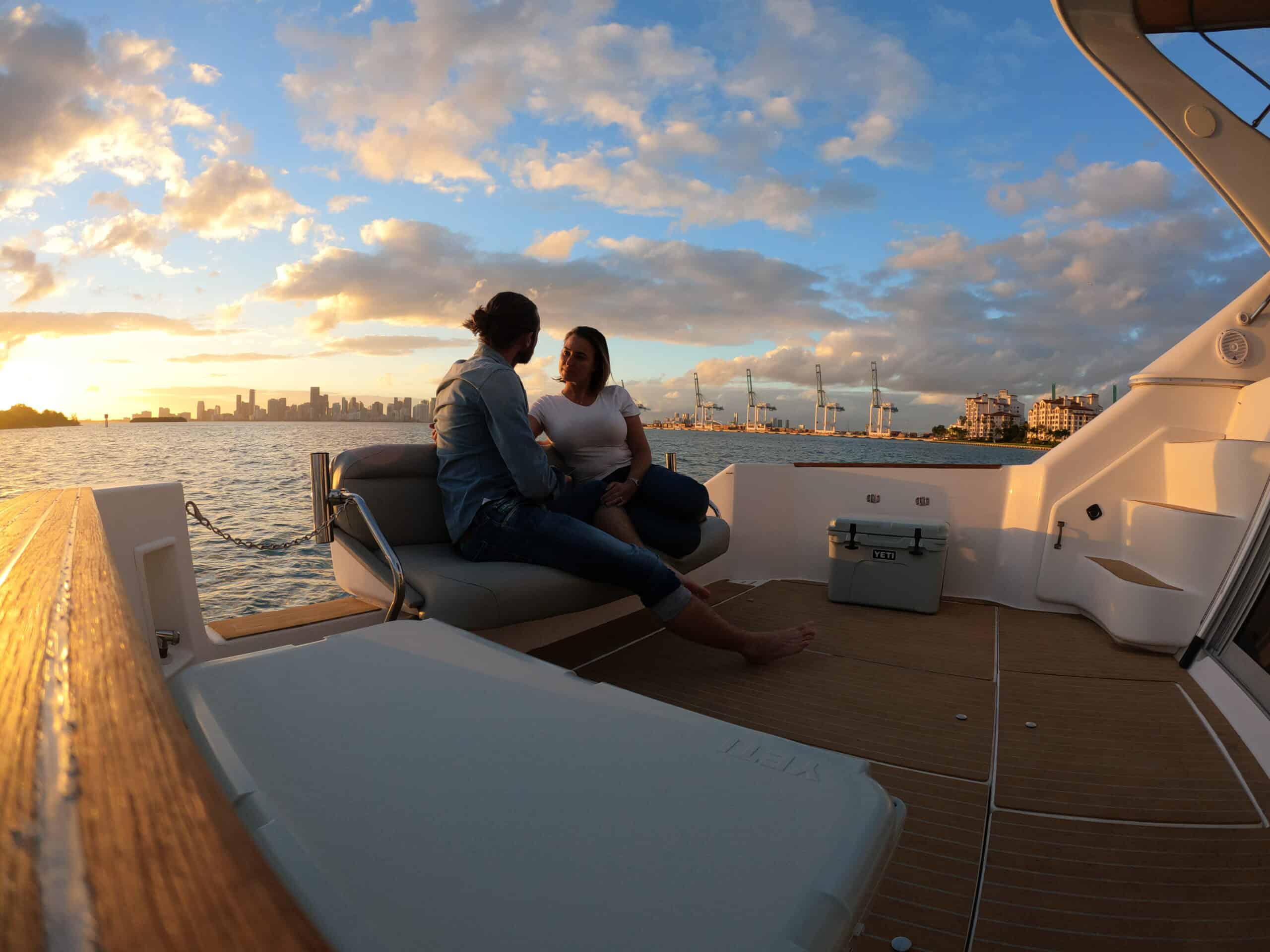 A couple sits facing each other on the stern of a boat during sunset, with the Miami skyline in the background.