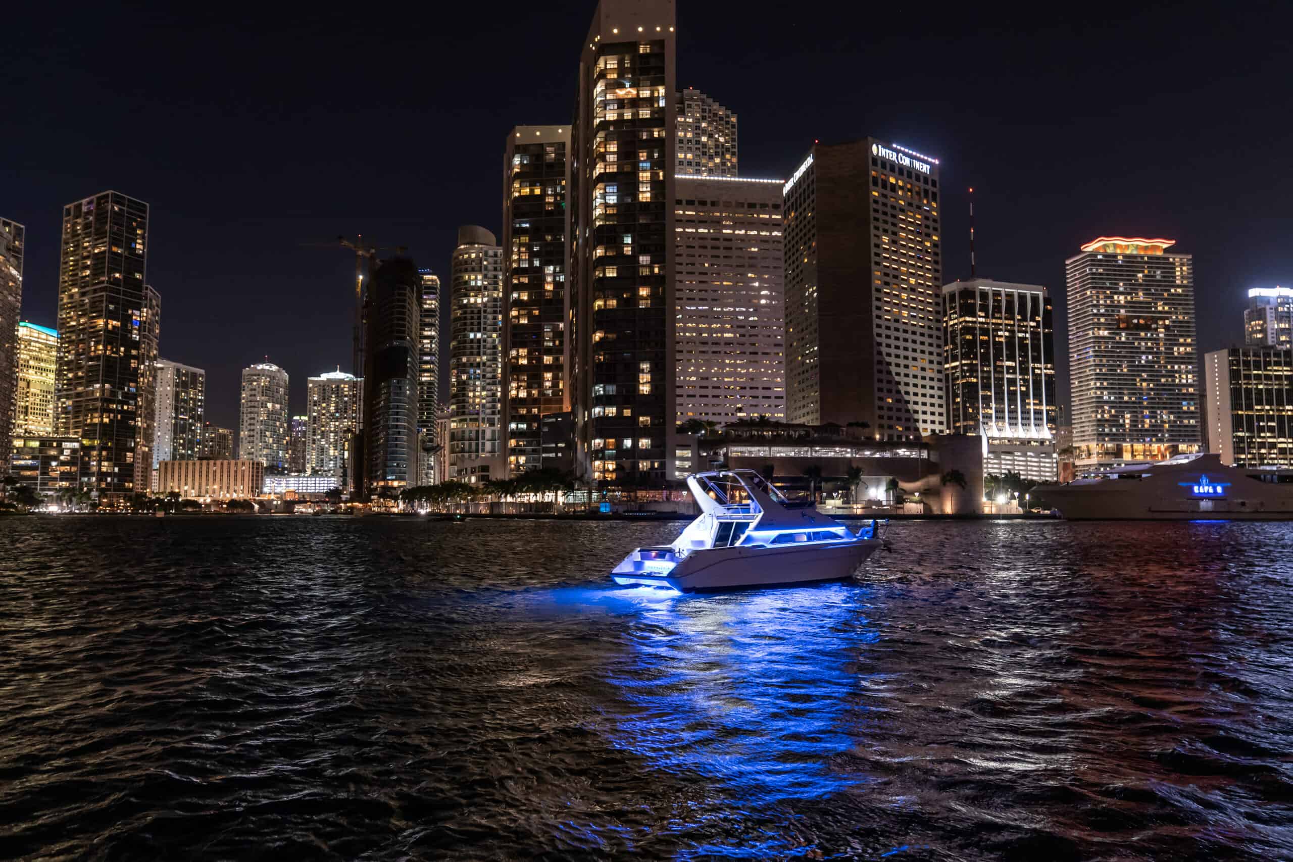 A boat with blue lights floats on a river in Miami at night, with a backdrop of a brightly lit city skyline and high-rise buildings.