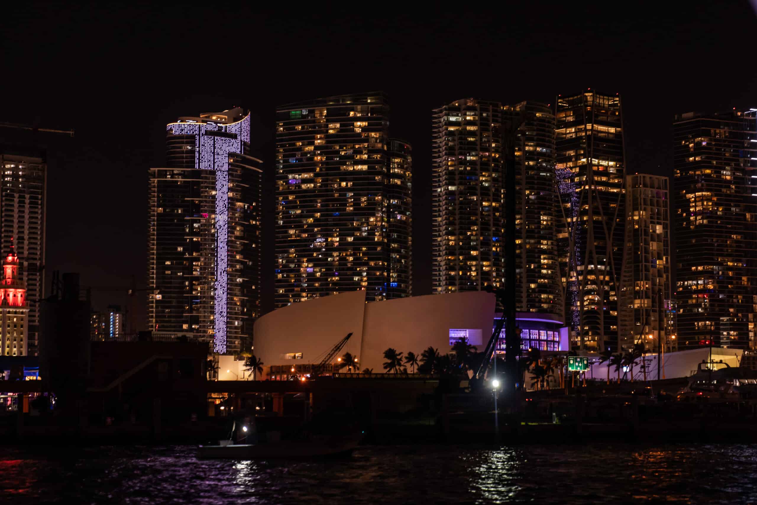 Night view of Miami's waterfront with illuminated skyscrapers and a boat on the river.