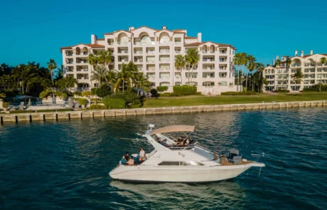 Aerial view of a white yacht with people onboard, cruising near a coastal luxury residential building in Miami under clear skies.