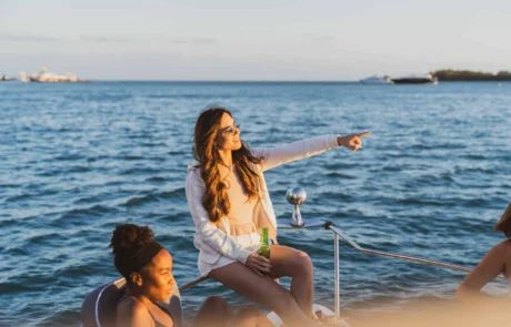 Three women enjoying time on a boat in Miami at sunset; one standing and pointing at the horizon, while the others sit, one holding a drink. Three women enjoying time on a boat in Miami at sunset; one standing and pointing at the horizon, while the others sit, one holding a drink.