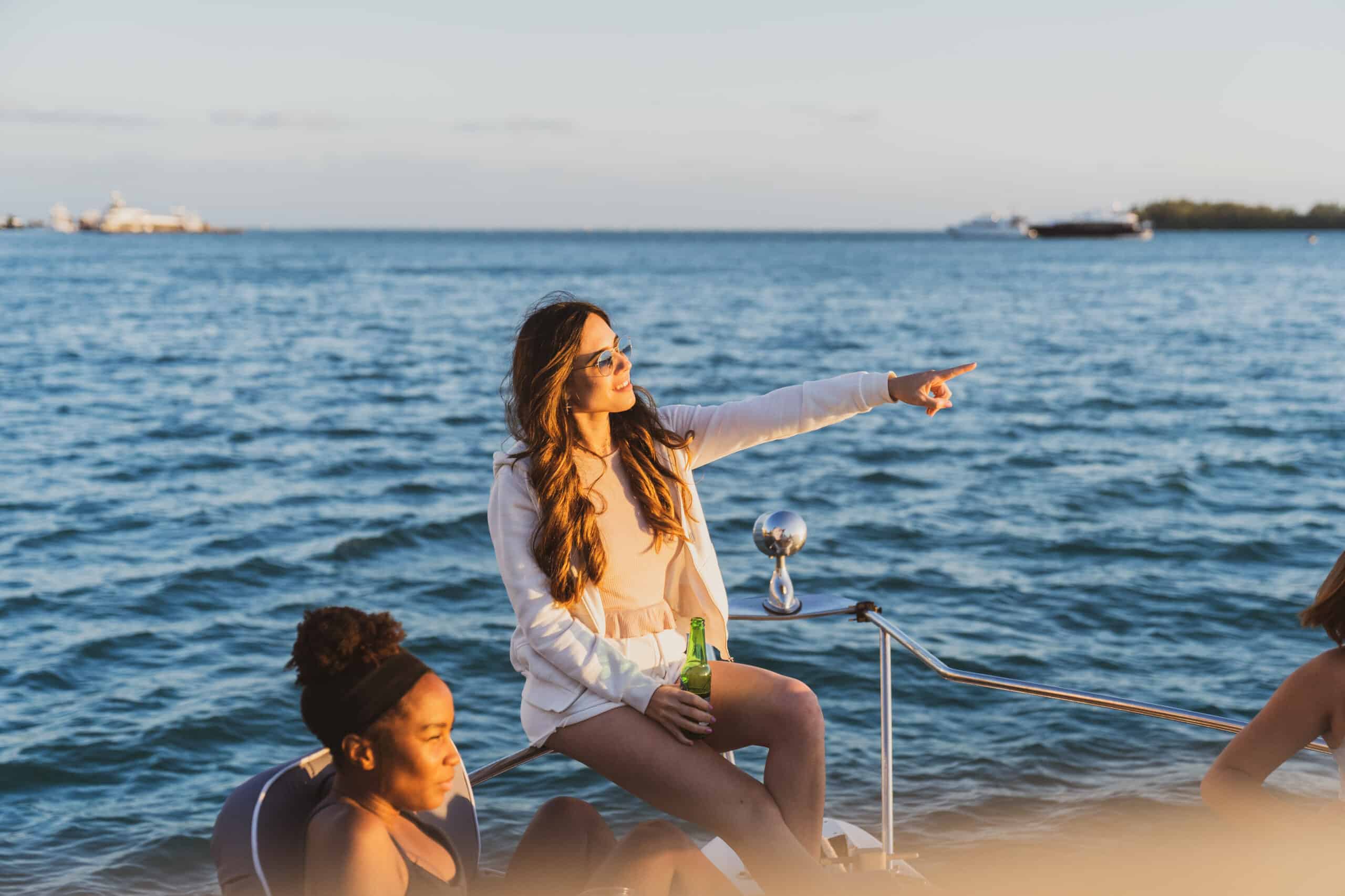 Three women enjoying time on a boat in Miami at sunset; one standing and pointing at the horizon, while the others sit, one holding a drink.