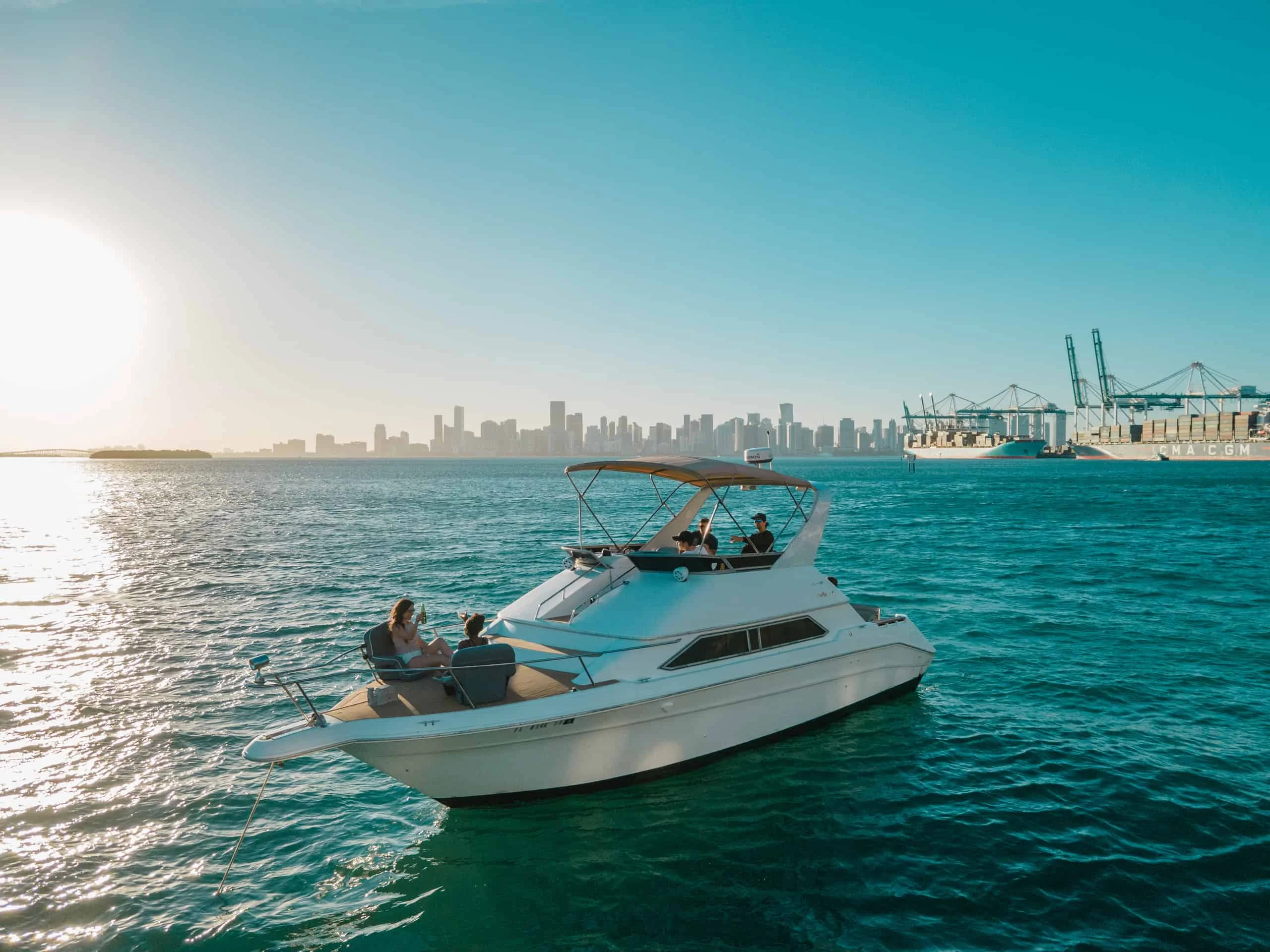 A small boat with two passengers cruising near the Miami skyline and port cranes under a clear blue sky. A small boat with two passengers cruising near the Miami skyline and port cranes under a clear blue sky.