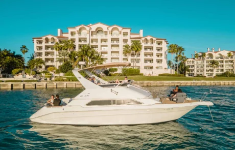 A white boat with several passengers cruising past a coastal apartment building in Miami under a clear blue sky.