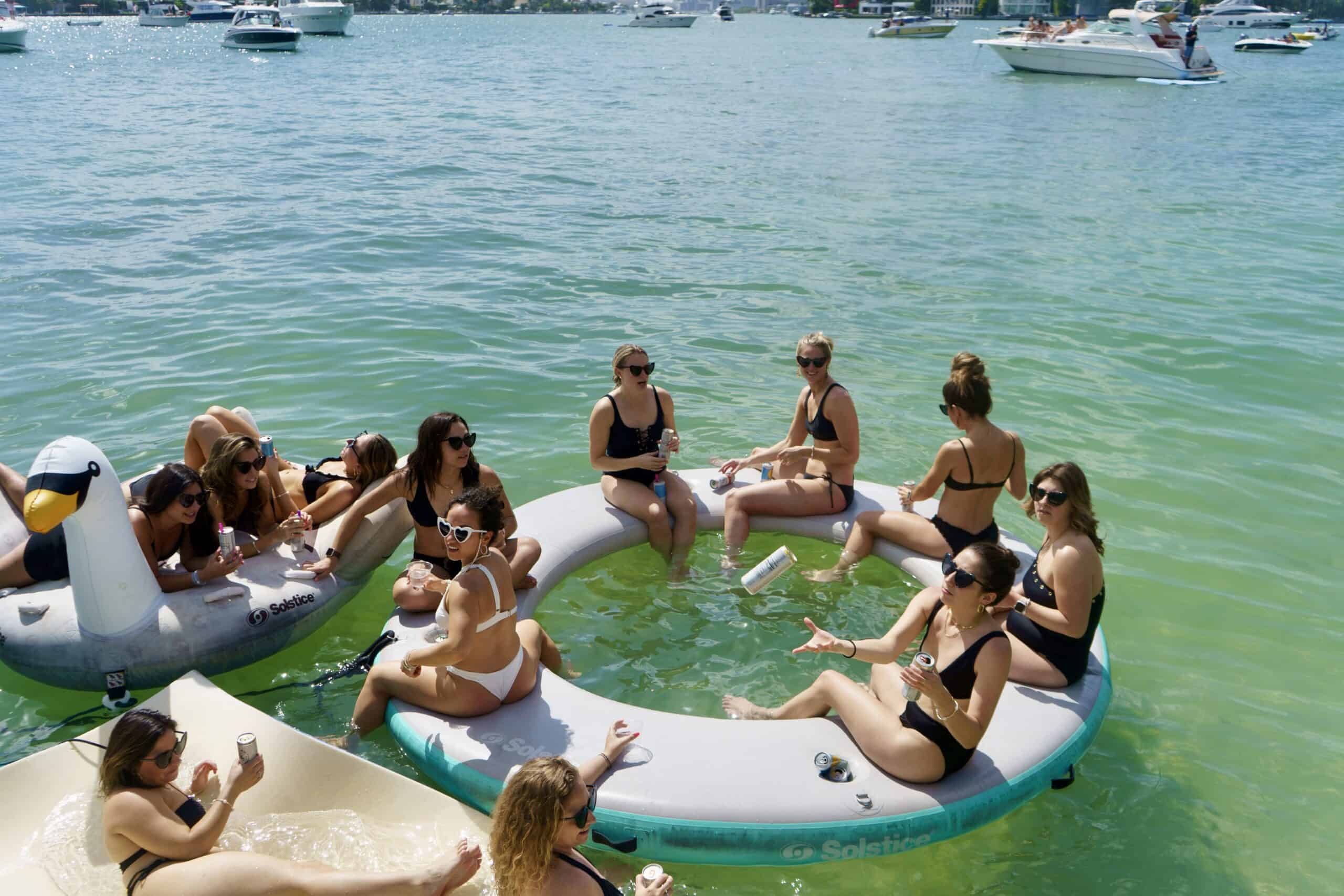 Group of people lounging on a large floating raft in a lake, surrounded by boats on a sunny day.