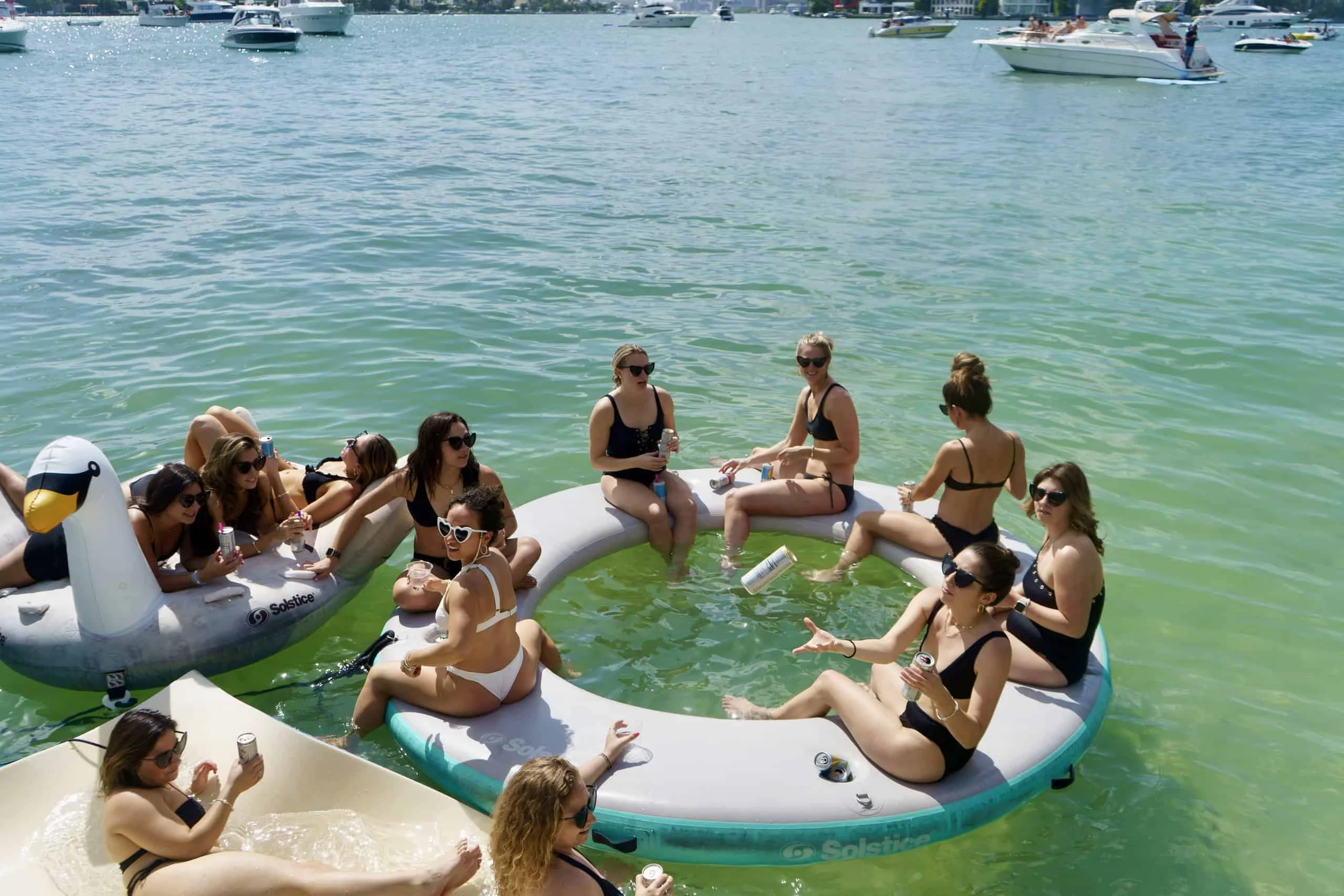 A group of women in swimsuits sitting on inflatable rafts, socializing on a sunny day on a calm, clear body of water in Miami, surrounded by boats.