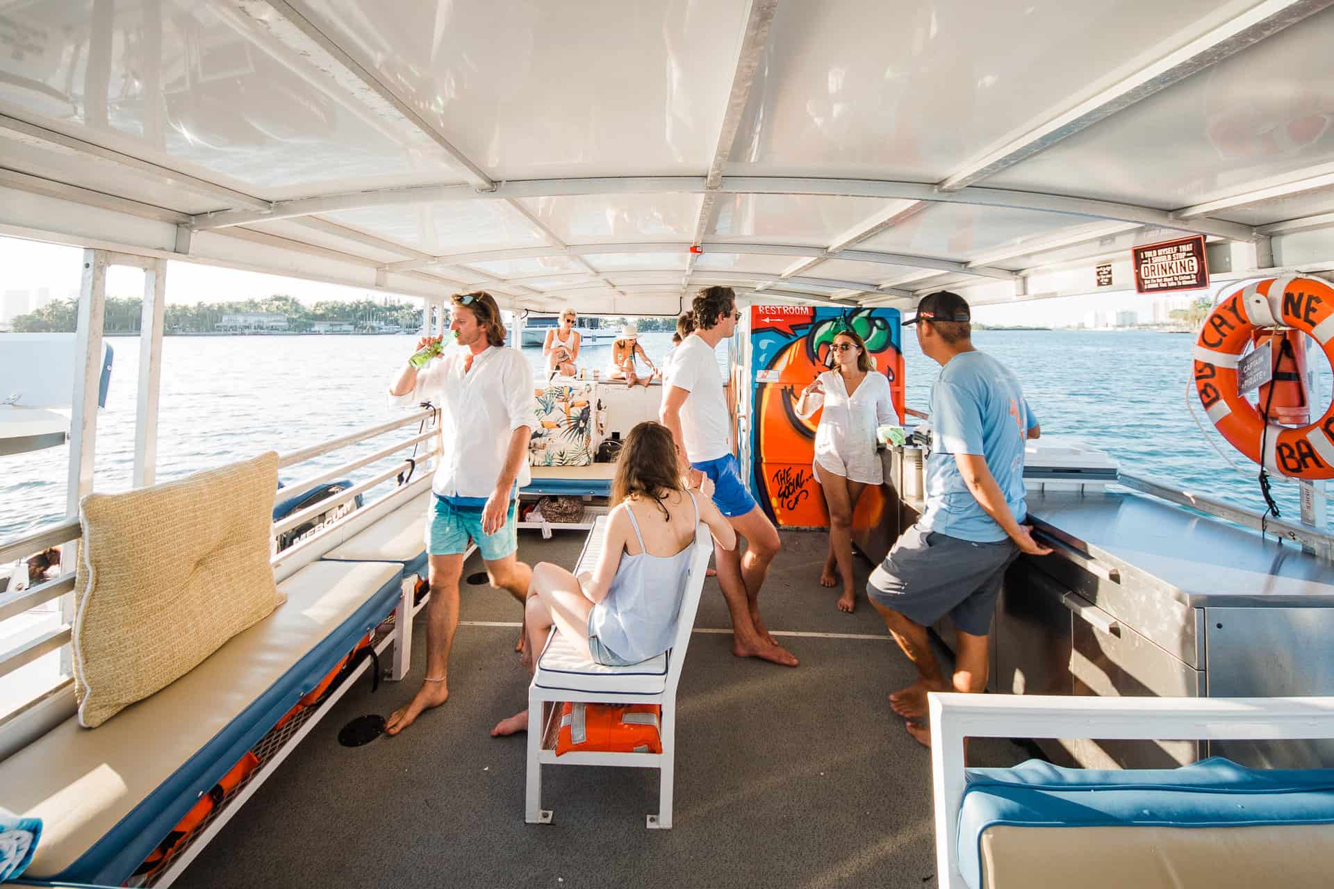 Group of people enjoying a boat ride in Miami on a sunny day, with some standing and others sitting, talking and drinking refreshments.