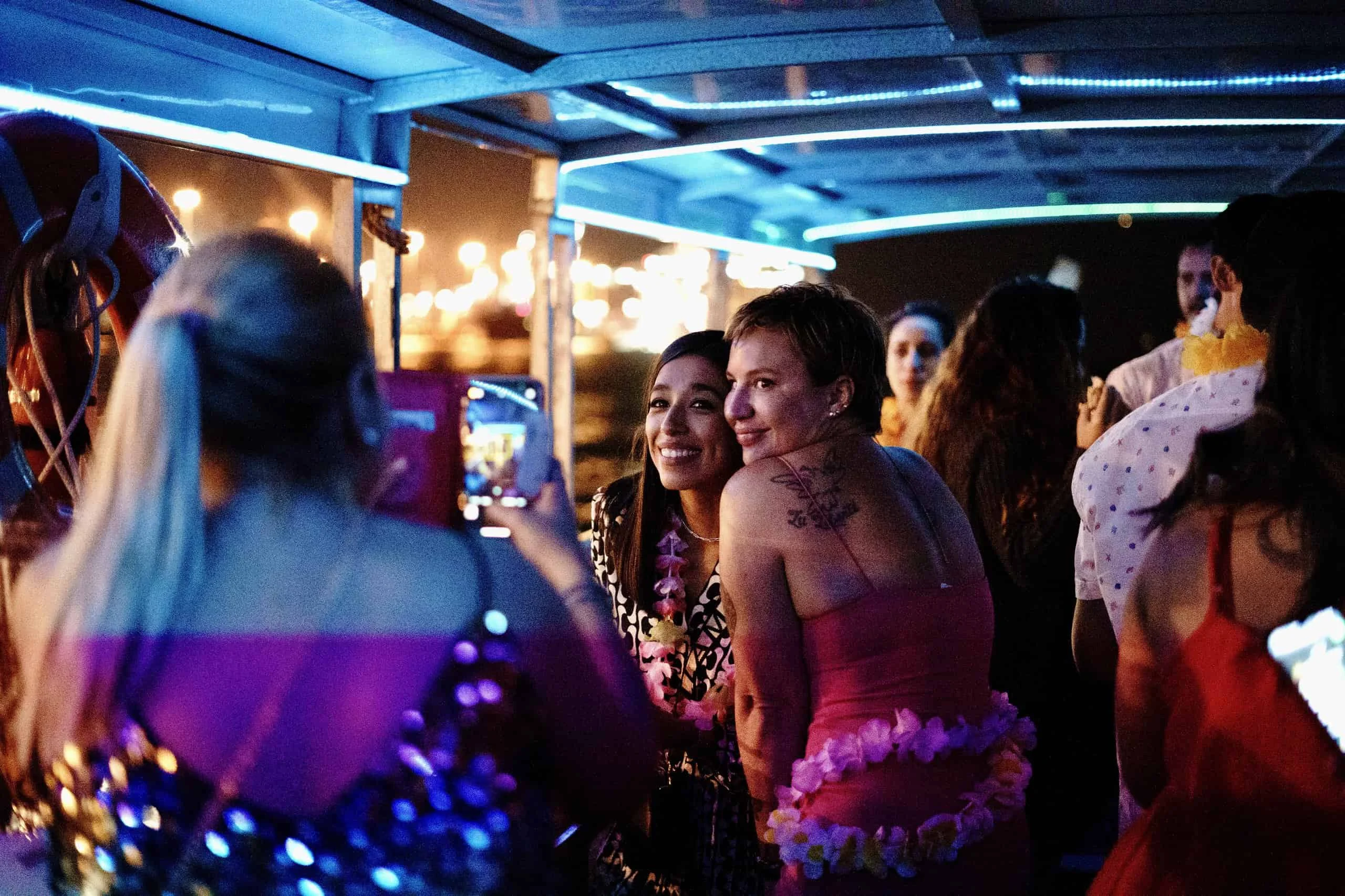 Two women smiling and taking a selfie on a boat at a vibrant costume party in Miami, with other guests and colorful lights in the background.