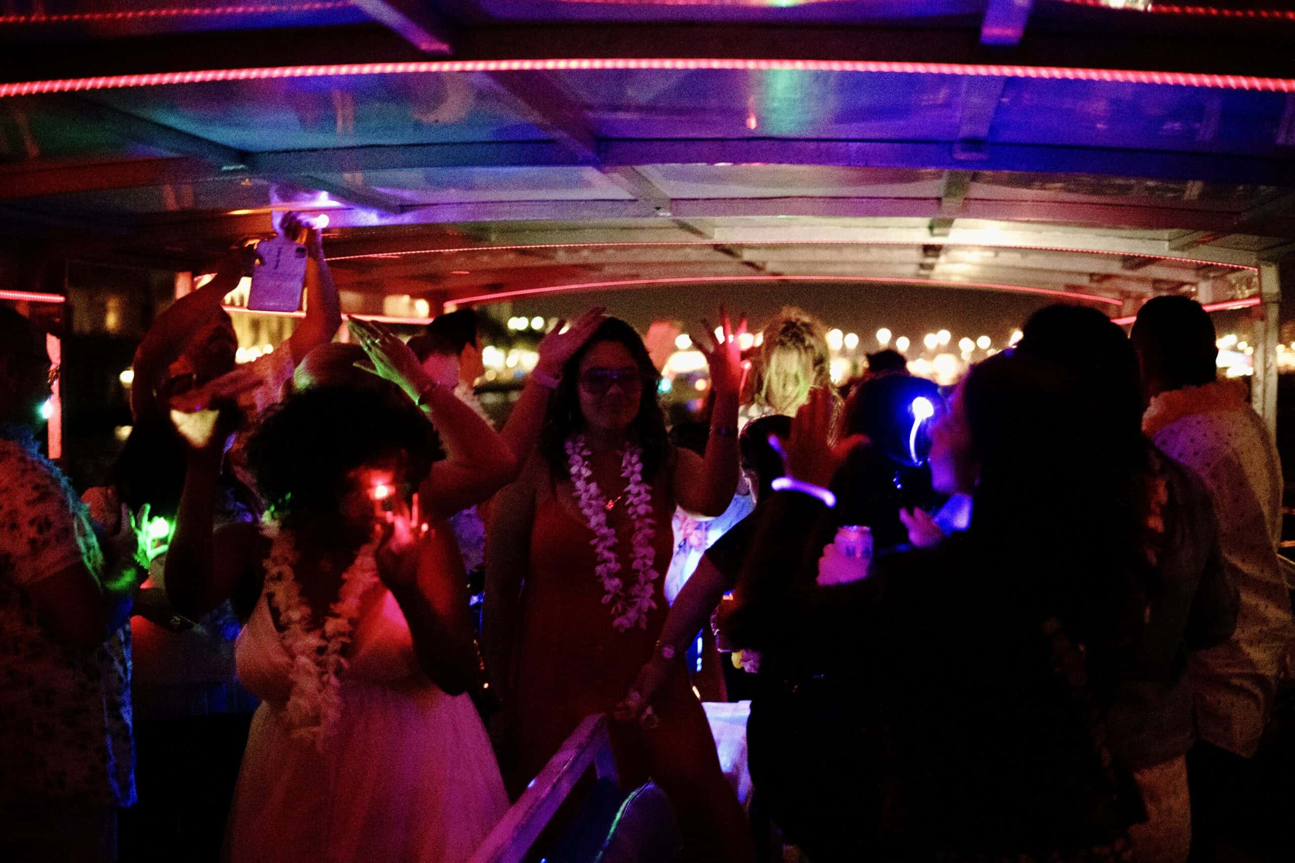 People dancing at a nighttime party on a Miami boat, decorated with colorful lights, wearing casual outfits and flower leis.