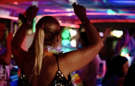 A woman dances at a vibrant boat party in Miami with colorful lights in the background, wearing a sequined top.