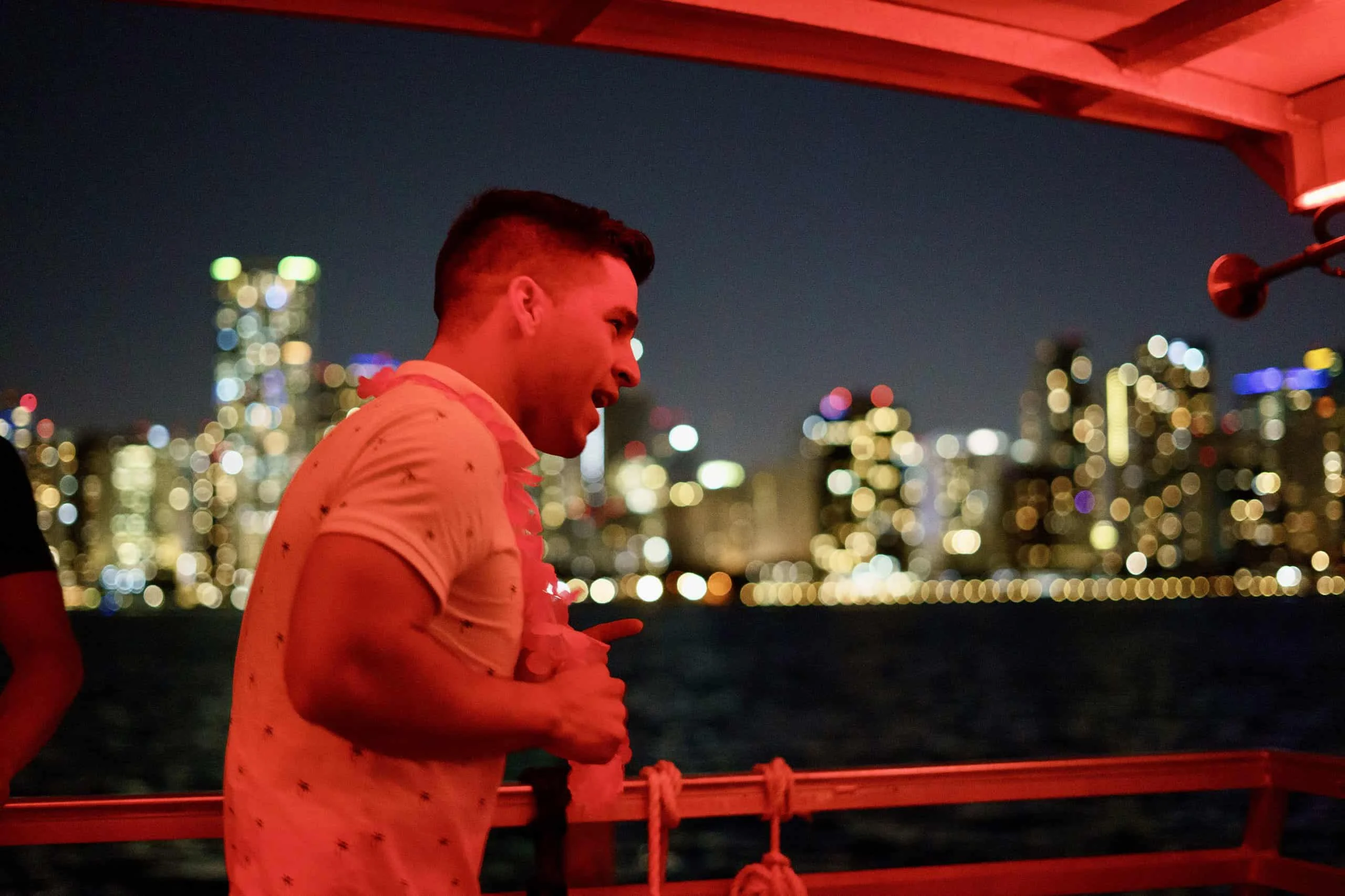 A man in a patterned shirt standing on a boat in Miami at night, with a brightly lit city skyline in the background. A man in a patterned shirt standing on a boat in Miami at night, with a brightly lit city skyline in the background.