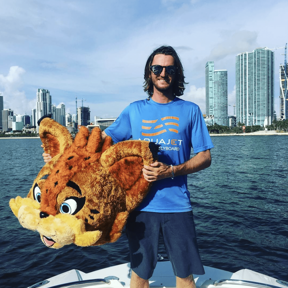 Man with sunglasses and a beard holding a large lion mascot head on a boat in Miami, with a city skyline in the background.