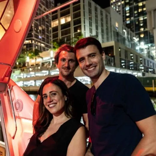 Three friends posing for a photo at night in an urban setting, with city lights in the background. Two men and one woman, all smiling at the camera in Miami.