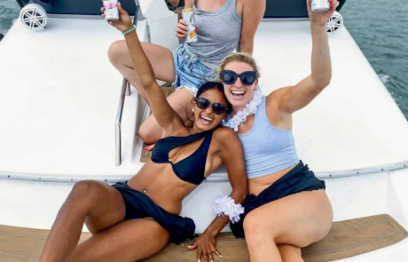 Three women holding drinks and smiling on the bow of a boat in Miami with the city skyline in the background.