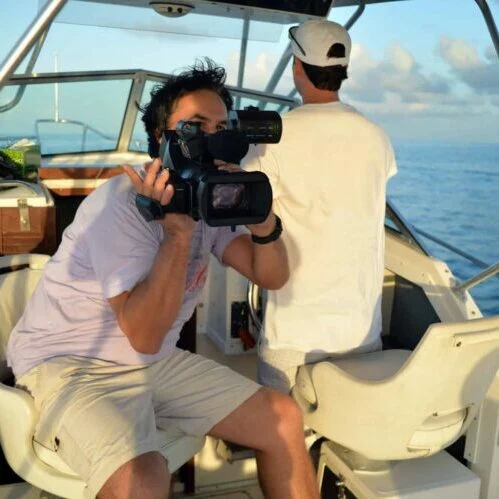 A man holding a video camera films from a boat in Miami, focusing into the lens, while another man looks out to sea.