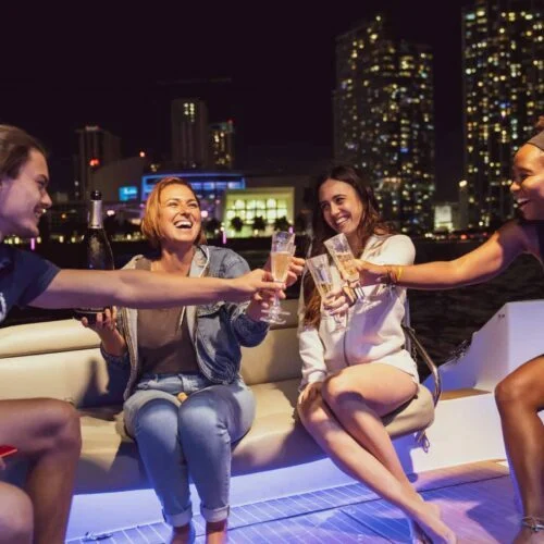 Four women toasting drinks on a boat in Miami at night with city lights in the background.