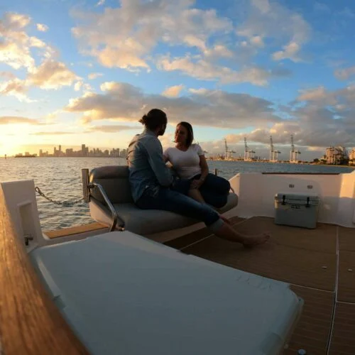 A couple sits on a boat in Miami, engaging in conversation at sunset with a city skyline and cranes in the background.