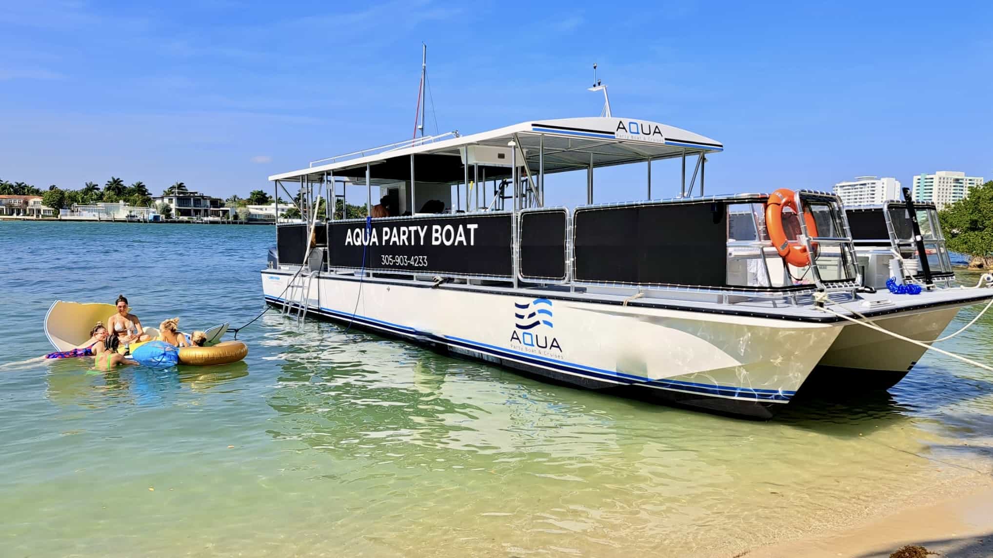 A large party boat named "Aqua" anchored near a Miami beach with people lounging on inflatable floats in clear blue water.