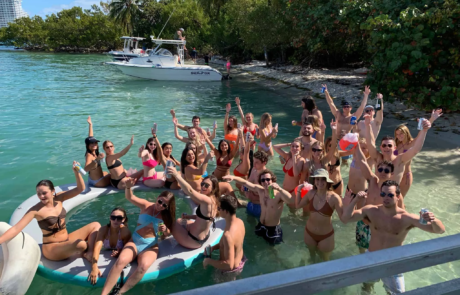 Group of people in swimsuits cheering and posing on a floating platform by a tropical Miami beach with boats nearby.