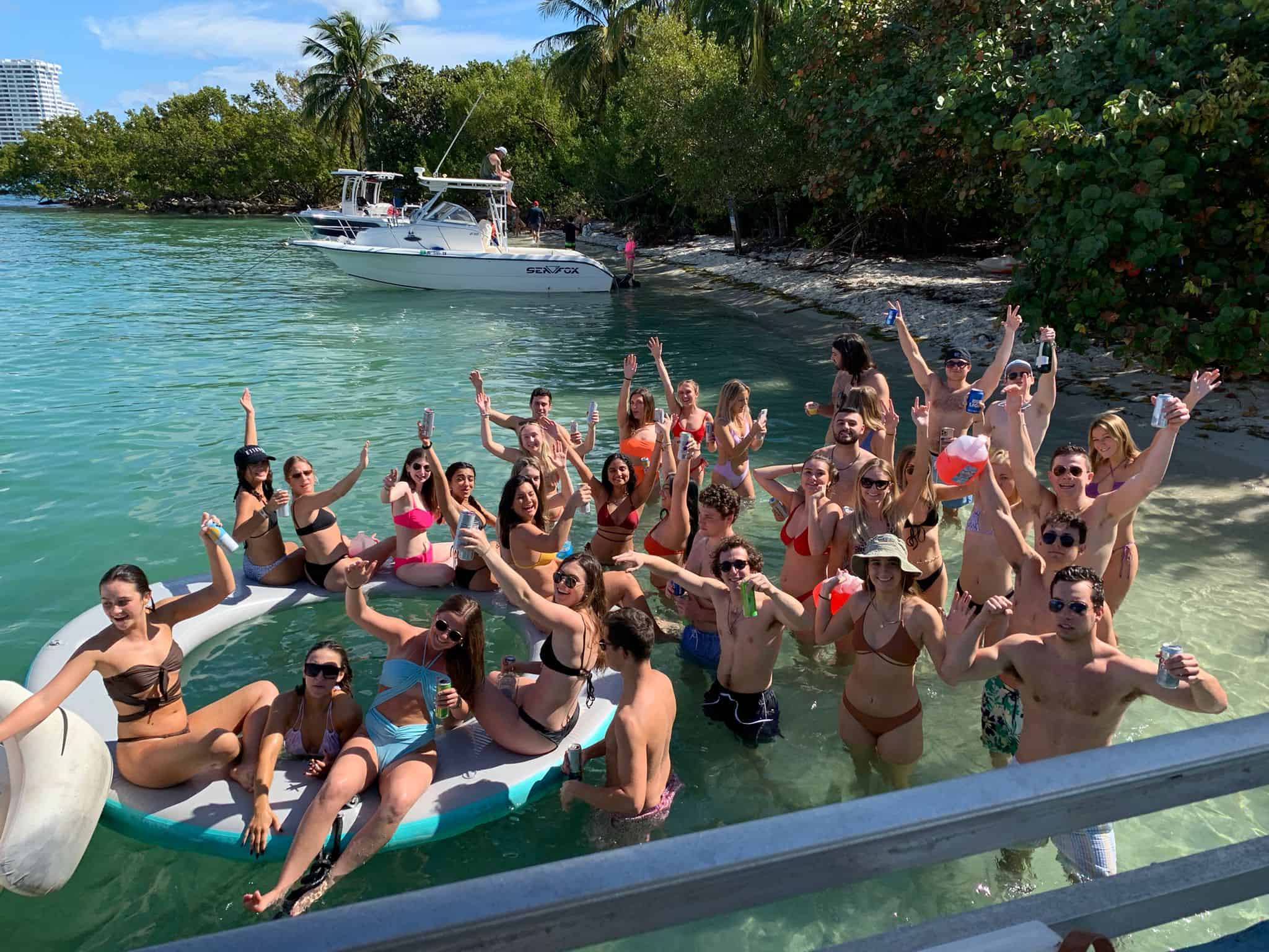 Group of people in swimsuits cheering and posing on a floating platform by a tropical Miami beach with boats nearby.