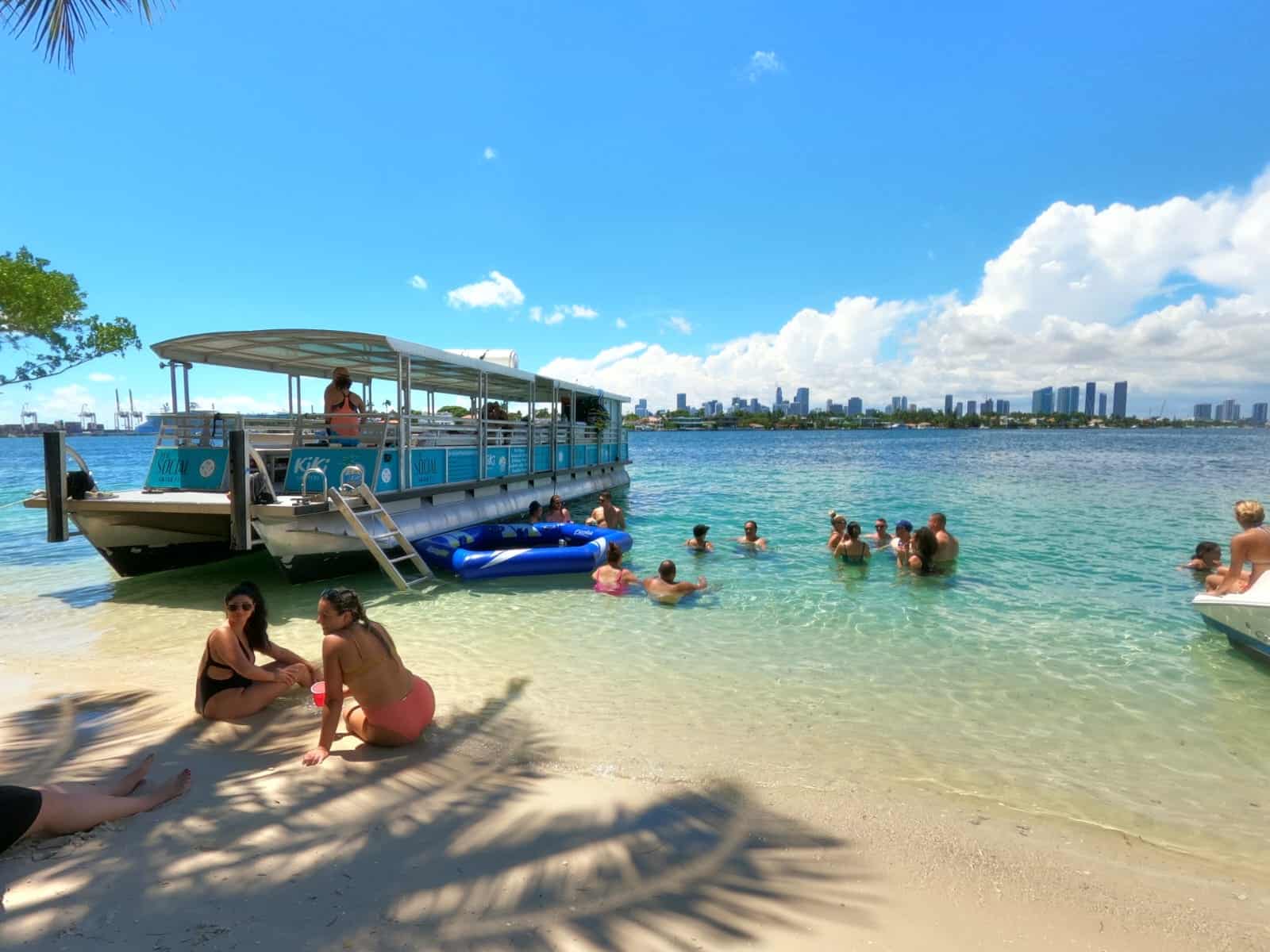 People enjoying a sunny day at a Miami beach with a city skyline in the distance and a boat moored near the shore.