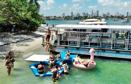 Aerial view of people enjoying a party on floating rafts near a boat in a waterscape, with the Miami skyline in the background.