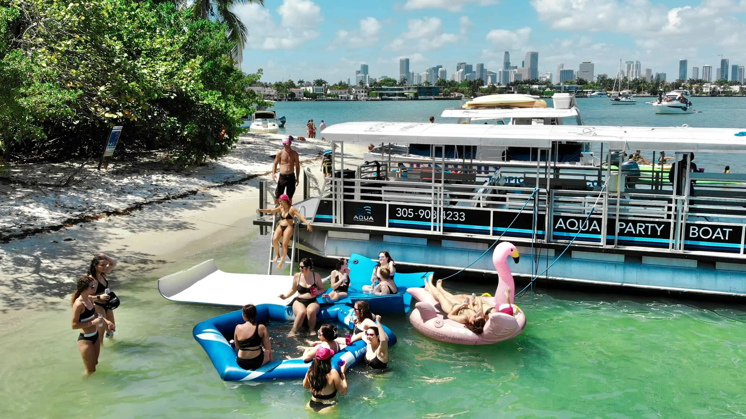 Aerial view of people enjoying a party on floating rafts near a boat in a waterscape, with the Miami skyline in the background. Aerial view of people enjoying a party on floating rafts near a boat in a waterscape, with the Miami skyline in the background.