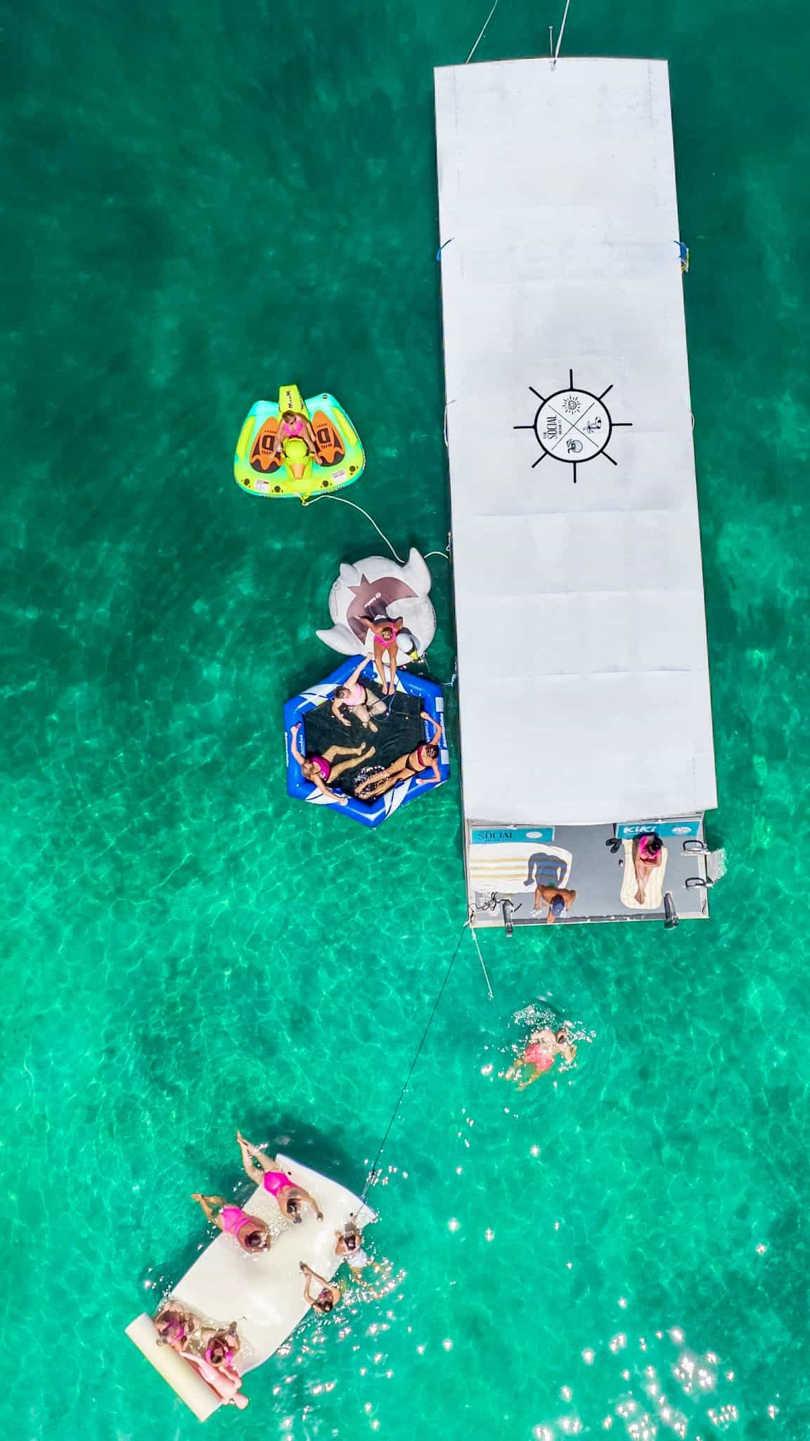 Aerial view of a boat surrounded by floating mats and people swimming in clear turquoise water near Miami.