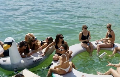 Group of people enjoying a sunny day on large inflatable floaties in water, featuring a swan and a unicorn, with boats in the background off the coast of Miami.