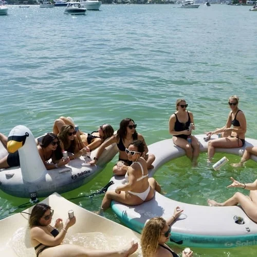 Group of people enjoying a sunny day on large inflatable floaties in water, featuring a swan and a unicorn, with boats in the background off the coast of Miami.