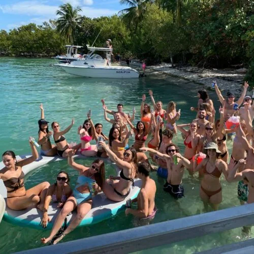 A group of young people in swimwear cheerfully celebrating in the water near a boat off the Miami coast, raising their arms and holding drinks.