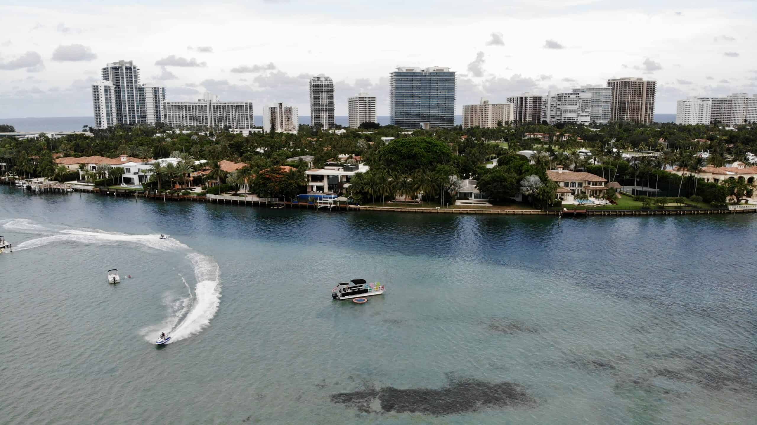 Aerial view of Miami's coastal area with luxury homes, high rises in the background, and boats on clear blue water.