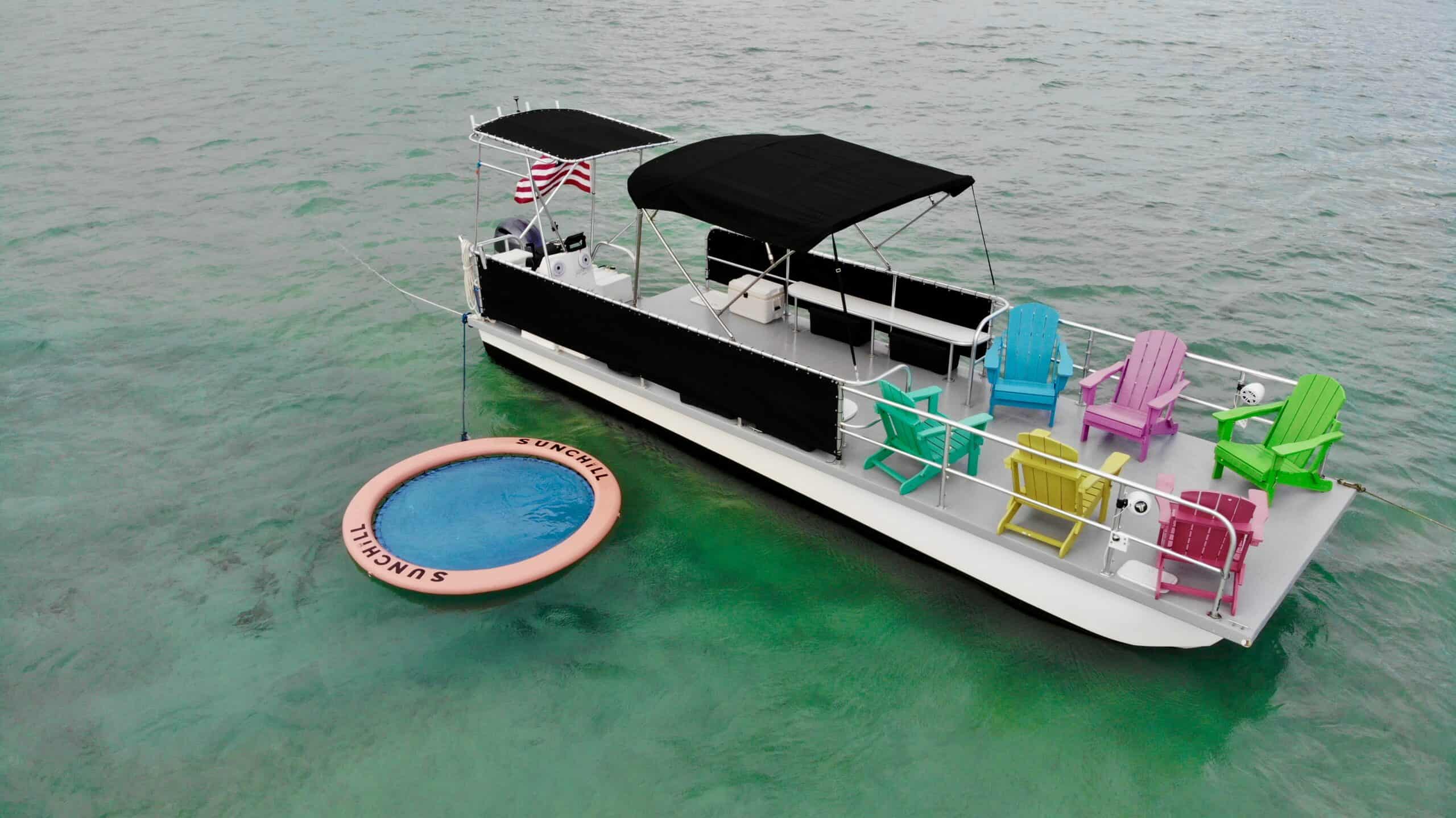 Aerial view of a colorful party boat with bright chairs floating in clear, greenish water near Miami, accompanied by a large pink inner tube marked "Summerville.