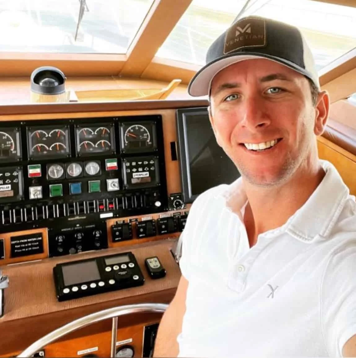 Man in a white polo and cap smiling at the camera in a boat's cockpit off the coast of Miami, with various navigation instruments visible.