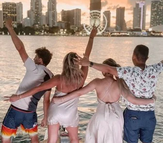 Four friends holding hands in the air on a boat in Miami, enjoying a sunset with a city skyline in the background.