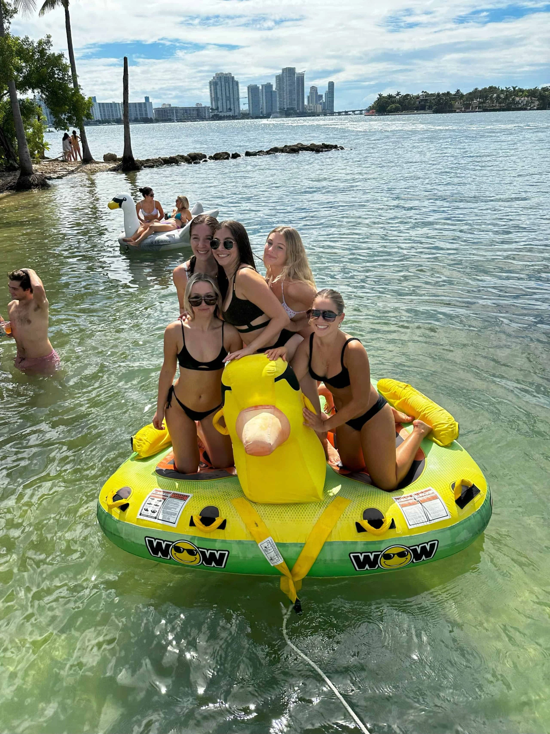 Group of friends enjoying a sunny day on a large yellow rubber duck float in calm waters with Miami’s skyline in the background. Group of friends enjoying a sunny day on a large yellow rubber duck float in calm waters with Miami's skyline in the background.
