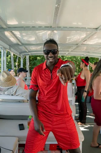 Man in a red outfit pointing at the camera, smiling, with others in the background on a sunlit boat in Miami. Man in a red outfit pointing at the camera, smiling, with others in the background on a sunlit boat in Miami.