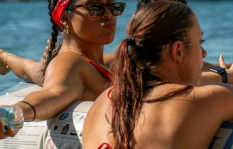 Two women in swimwear and sunglasses enjoying a sunny day on a boat in Miami, one wearing a red headband and pouting at the camera.