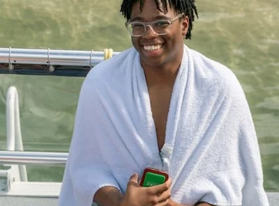 A young man wearing glasses and green shorts sits on a boat in Miami, wrapped in a white towel, smiling while holding a green phone.
