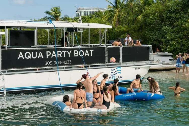People in swimsuits enjoy a sunny day on and around a floating inflatable raft near the “Miami party boat” anchored by a lakeside. People in swimsuits enjoy a sunny day on and around a floating inflatable raft near the "Miami party boat" anchored by a lakeside.