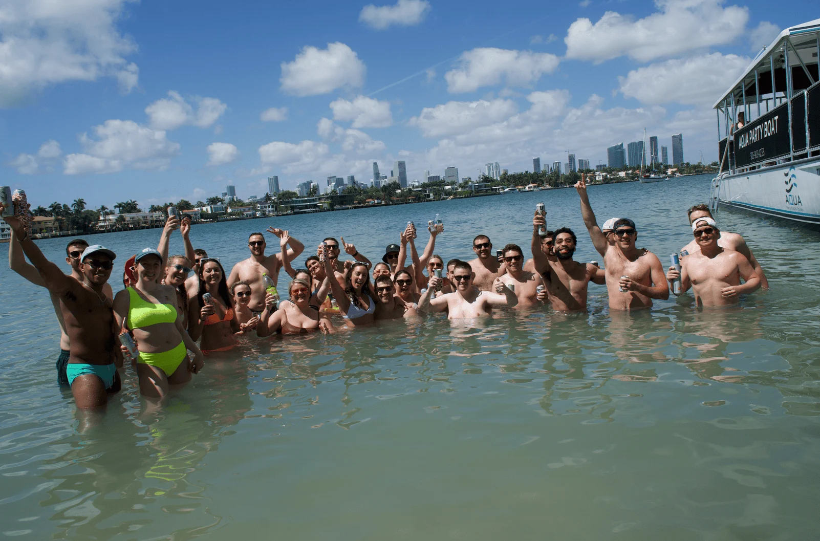 A group of people standing in shallow water near a boat in Miami, raising their drinks and smiling, with a city skyline in the background. A group of people standing in shallow water near a boat in Miami, raising their drinks and smiling, with a city skyline in the background.